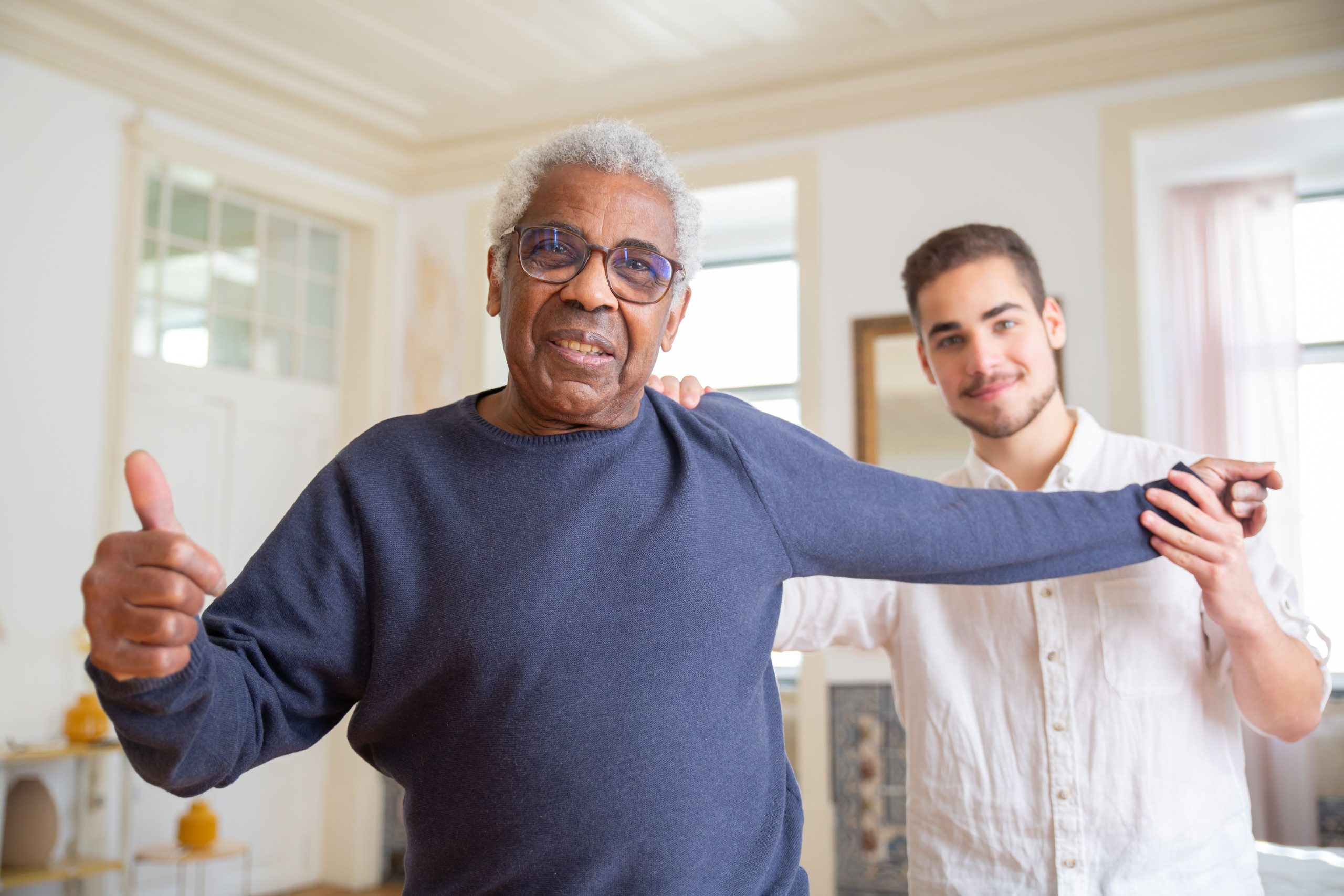 A senior man doing arm extension exercises with a young male instructor or caregiver while giving a thumbs-up to the camera.